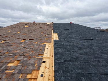 Wide environmental shot of a Sioux City suburban home mid-replacement, old shingles stripped to bare decking on one half, new dark shingles installed on the other half, overcast Midwest sky, workers visible at the ridge