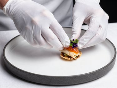 Extreme close-up of hands in white gloves placing a garnish on a plated appetizer, bright directional studio light, charcoal-rimmed plate on a white linen surface, tight framing
