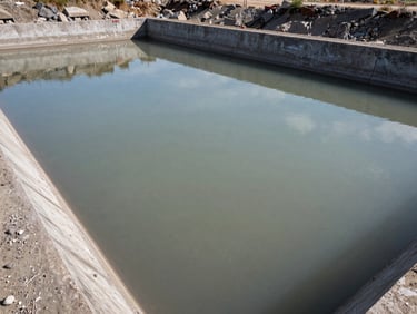 Mining site water treatment settling pond, grey sediment-laden water in a wide concrete basin, overhead medium shot showing water clarity gradient from inlet to outlet under open sky