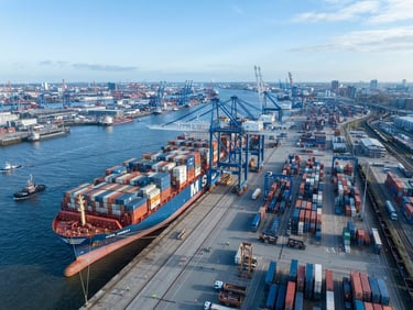 Massive container ship docked at a modern industrial port during bright daylight, cool blue maritime tones, high-angle editorial framing showing shipping containers and crane logistics, professional maritime photography.