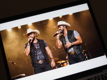Close-up of a smartphone or laptop screen showing a YouTube video thumbnail of two sertanejo performers on stage, warm stage lighting, gold tones, dramatic shadow, concert venue visible in background
