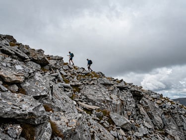 Wide environmental shot, two hikers seen from a distance traversing an exposed rocky ridgeline, overcast sky, dramatic drop on one side, natural flat Balkan mountain light, no enhancement