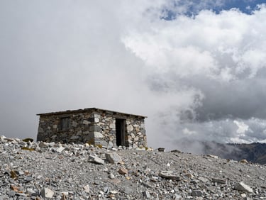 Exterior of a low stone refuge shelter on a bare alpine ridge, grey scree foreground, heavy cloud cover rolling in from the right, no figures, raw and exposed atmosphere, documentary framing