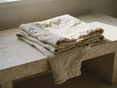 Soft folded natural linen fabric draped over a minimalist stone bench, warm morning light, gentle shadows, 35mm film grain.