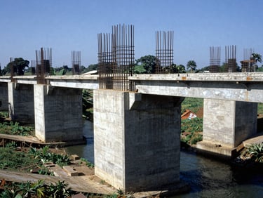 Wide environmental shot of a concrete bridge foundation built over a river canal in a Colombian agricultural region, steel reinforcements visible, bright daylight.