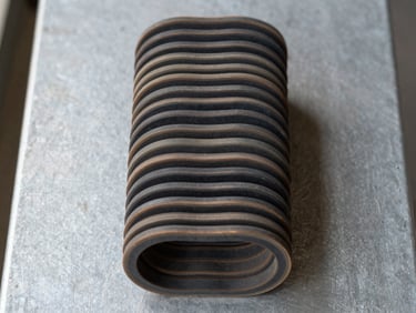 Overhead flat-lay of die-cut rubber gaskets stacked in a neat column on a gray steel surface, showing cross-section thickness, natural workshop daylight