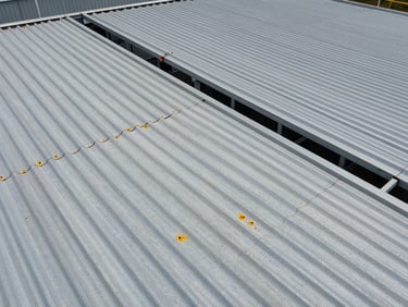 Close-up aerial drone view of corrugated industrial roof panels with visible corrosion spots under overcast daylight, structural frame partially visible