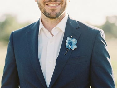 Candid portrait of a smiling man in a navy suit, soft golden-hour light, shallow depth of field, delicate hand-painted blue watercolor floral accents.