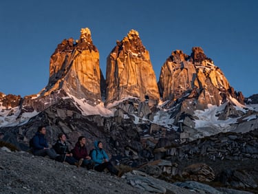 Wide shot at dusk looking across the Torres del Paine massif, a group of four hikers resting on rocky ground in the foreground left, golden light raking the granite towers, deep blue sky above