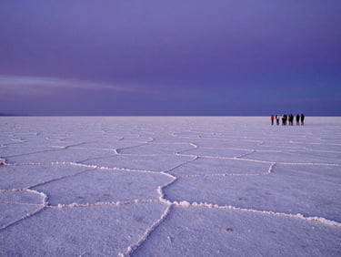 Wide environmental shot of the Atacama salt flats at blue hour, the horizon perfectly flat and infinite, a small cluster of travelers standing at the edge of the flats to the right, deep violet sky above