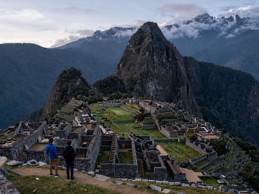 Panoramic early morning blue-hour view of Machu Picchu terraces with low cloud drifting through the valley below, two travelers standing quietly at a stone wall in the left third, soft ambient light