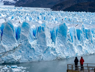Close wide-angle of the Perito Moreno glacier face at early morning, blue ice wall filling the frame, two travelers in cold-weather gear standing at the viewing platform lower right, mist rising off the water
