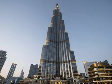 Straight-on dusk photograph of Downtown Dubai's Burj Khalifa tower base looking upward, facade lighting just activating, wide base composition showing plaza scale, no people foregrounded