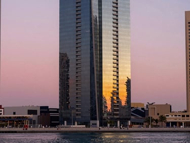 Straight-on architectural facade detail of a Dubai Marina tower at twilight, reflective glass panels catching pink and gold sky, shot from the marina boardwalk, water in the foreground