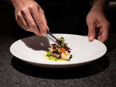 Hands of a chef plating a highly structured micro-seasonal dish with tweezers, dark editorial style, deep shadows