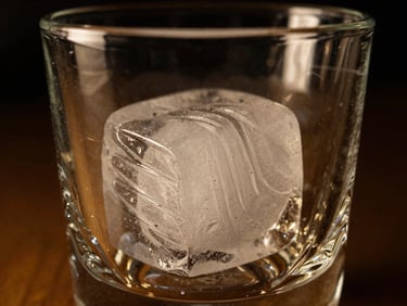 Close-up of a large, perfectly clear hand-carved ice cube in a crystal glass, dramatic low-key lighting, rich gold reflections, shallow depth of field