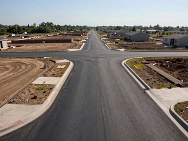 Wide midday photograph of a completed urban subdivision road network showing new asphalt pavement, kerbs, and stormwater drainage in a residential development, no workers, scale visible from surrounding unfinished lots