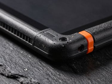 Extreme close-up of a rugged military tablet corner, reinforced rubber bumper, visible dust and water droplets, dramatic side-lighting, dark slate background, 35mm