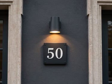 Straight-on view of a modern house number plate illuminated by a warm downward sconce on a dark charcoal wall