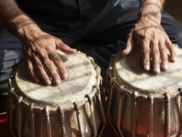 Overhead close-up of two hands on a tabla pair, fingers mid-stroke on the bayan, natural window light casting short shadows across the drum skin