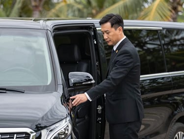 Close-up of a professional chauffeur opening the door of a pristine black executive van, tropical foliage softly blurred in the background, sharp daylight.