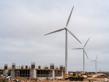 Ground-level documentary photograph of a wind turbine base under construction in an open Castilian plateau, heavy equipment and concrete foundation formwork visible in the foreground, overcast diffuse light, no people in frame, technical site survey context