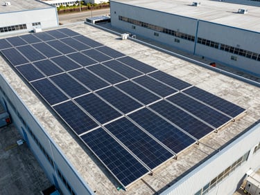 Overhead drone shot of a rooftop solar array on an industrial warehouse, panels arranged in tight rows across a flat concrete roof, harsh midday light casting minimal shadows, neutral urban context