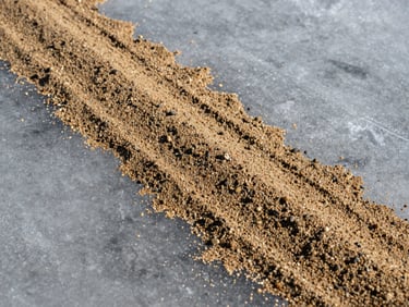 Close-up of sand and fine gravel abrasive mixture spread on icy pavement surface, grit particles visible against grey ice under flat winter light, texture-forward framing