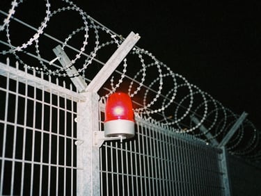 Close-up of security infrastructure, metal fence with barbed wire against a dark night sky, desaturated tones, red warning light.
