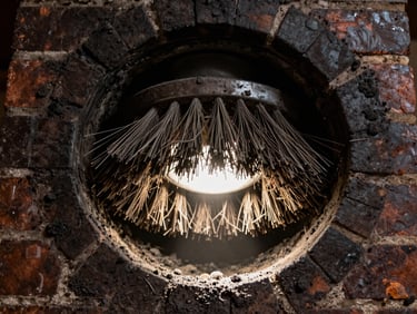 A close-up view inside a dark brick chimney flue, with heavy metal brush bristles scrubbing away soot, illuminated by a sharp beam of light from below.