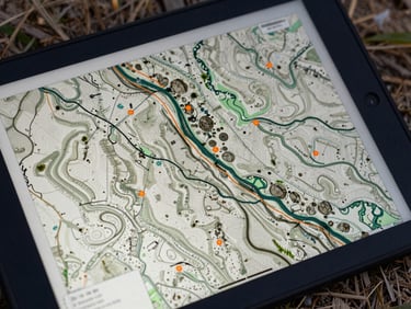 Close-up of a rugged field tablet displaying a complex GIS topographic map overlay with orange coordinates, natural outdoor lighting.