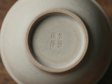 Overhead close-up of a maker's mark on the base of a ceramic bowl, diffuse daylight, muted warm tones, fine surface texture visible