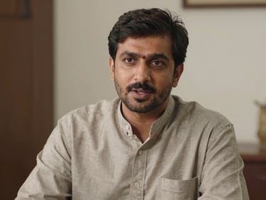 Medium shot of a Pakistani man in his late thirties speaking directly to camera with natural confidence, natural interview lighting, authentic expression, no studio setup, plain indoor background