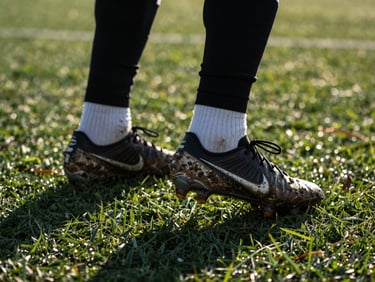 Tight close-up of muddy football boots on wet green turf during an intense training session, sharp evening floodlight casting long shadows