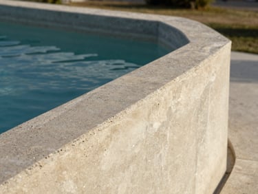 Close-up north-daylight shot of a cast-in-place above-ground concrete pool edge, the aggregate finish in warm graphite and cream, water surface catching diffuse light, pool wall rising as a sculptural parapet, surrounding landscape softly out of focus