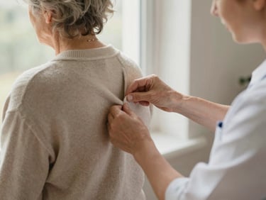 A caregiver gently helping a senior woman adjust her warm cardigan by a sunlit window, focusing on hands and a supportive, respectful interaction.