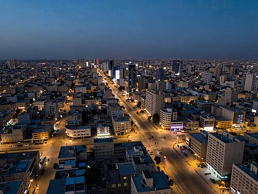 Aerial perspective of a modern Iraqi city district at dusk, LED street lighting grid visible from above, infrastructure corridors cutting through the urban fabric, deep blue sky at the horizon
