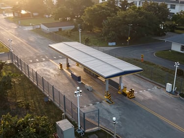 Overhead aerial perspective of a border station facility at dawn, perimeter fencing with integrated camera towers, structured access lanes, hard directional sunrise light from the east, no people