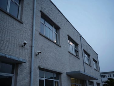 Hospital facility exterior at dusk, security camera mounts visible on facade, cool practical lighting, wide architectural framing, no people, documentary tone