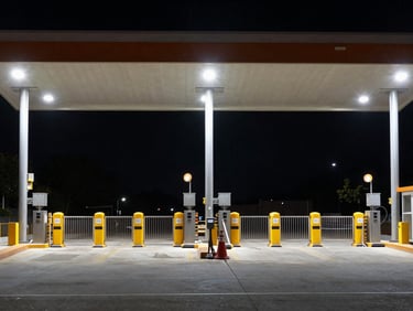 Border checkpoint infrastructure at night, tall lighting columns illuminating a wide gate structure, security hardware visible on posts, overhead framing, no personnel, high-contrast ambient light