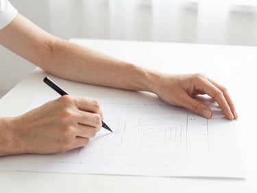 Bright high-key fashion photography, soft natural daylight, close-up of hands drafting a pattern on a clean white table, crisp details, 50mm lens