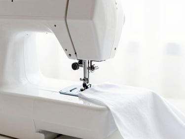 Bright high-key fashion photography, soft natural daylight, close-up of a modern sewing machine in motion with white fabric, crisp details, 50mm lens