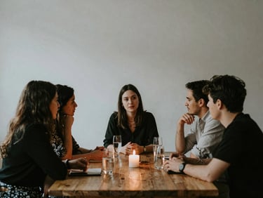 Candid medium shot of people talking around a candlelit wooden table at an evening event, low contrast, warm atmospheric shadows