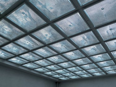 Wide architectural shot of a ceiling installation in progress showing a diffuser array of perforated metal panels in a grid pattern, cool north-facing construction light, empty commercial space, no people, detail of panel joints and fixing hardware visible