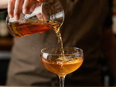 Close-up of hands pouring a perfectly clear amber liquid from a mixing glass into a coupe, shallow depth of field, warm editorial style