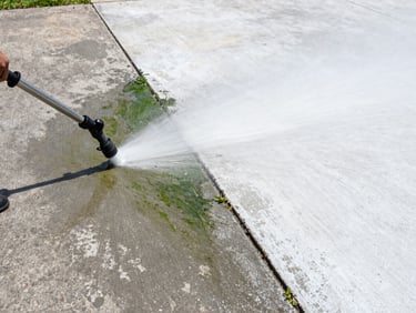 Ground-level wide shot of a concrete driveway being pressure-washed, visible split between dirty grey-green algae-stained concrete on the left and bright white clean concrete on the right, professional equipment nozzle visible, clear daylight, documentary