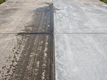 Close-up of a concrete driveway split down the center — left half covered in dark algae and grime, right half freshly pressure-washed to bright clean gray, sharp dividing line visible, bright Maryland daylight, overhead documentary framing