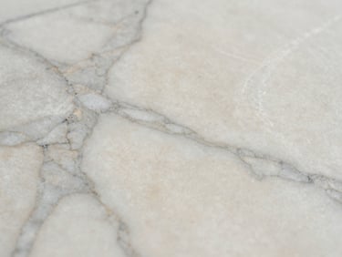 Extreme close-up of a honed marble slab surface, soft grey veining on warm white ground, photographed under diffused north-facing light showing matte finish texture, no staging