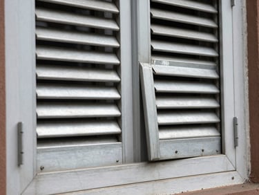 Close-up of two bent aluminium shutter slats on a residential window, one slat buckled outward, natural daylight, worn paint on surrounding frame visible