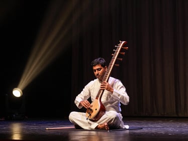 Wide environmental photograph of a sarangi player on a dimly lit concert stage, instrument held vertically against chest, warm copper spotlight from stage left, dark curtain behind, no audience in frame